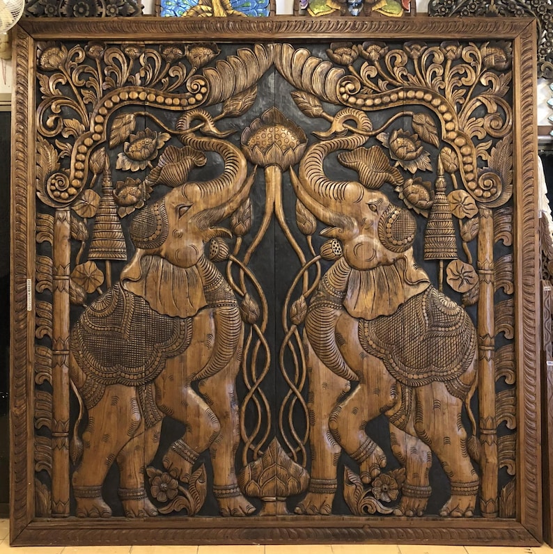 Puede incluir: Un gran panel de madera tallada con dos elefantes enfrentados. Los elefantes est&aacute;n rodeados de dise&ntilde;os florales y foliares detallados, con un fondo oscuro. El marco es de color marr&oacute;n oscuro.
