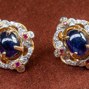 Puede incluir: Un par de pendientes de oro con una piedra preciosa azul rodeada de piedras blancas más pequeñas y acentos rojos.