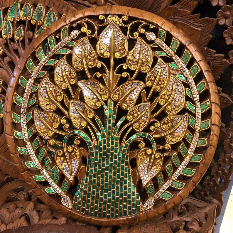 May include: A round wooden wall decor with a carved design of a tree with green and gold leaves. The tree is decorated with small, shiny, green and silver beads.