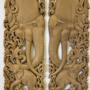 May include: A pair of intricately carved wooden wall panels featuring elephant heads with floral designs. The panels are made of light brown wood and have a traditional Thai style.