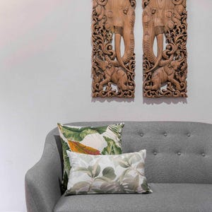 May include: Two intricately carved wooden panels depicting elephants with floral designs. The panels are hanging on a white wall above a grey sofa with two green and white patterned throw pillows.
