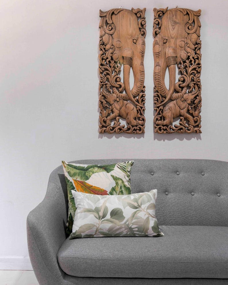 May include: Two intricately carved wooden panels depicting elephants with floral designs. The panels are hanging on a white wall above a grey sofa with two green and white patterned throw pillows.