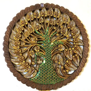 May include: A round wooden wall decor with a carved tree design. The tree is decorated with gold and green mosaic tiles. The background is a dark brown wood.