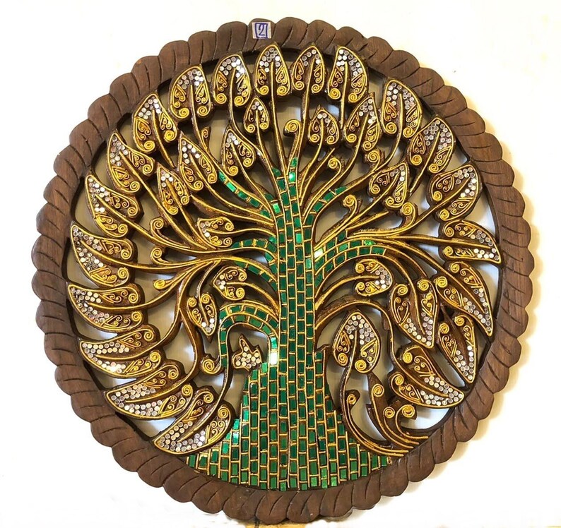 May include: A round wooden wall decor with a carved tree design. The tree is decorated with gold and green mosaic tiles. The background is a dark brown wood.