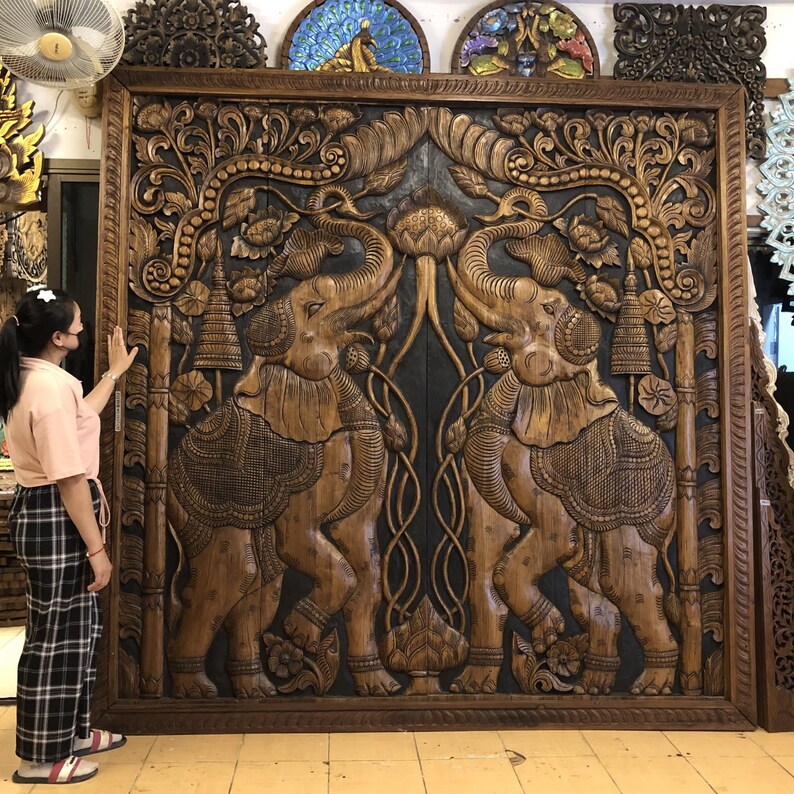 Puede incluir: Gran puerta de madera tallada con dos elefantes enfrentados. Los elefantes est&aacute;n detallados con patrones ornamentados y se alzan en medio de dise&ntilde;os florales y de hojas. La puerta est&aacute; enmarcada en madera oscura.