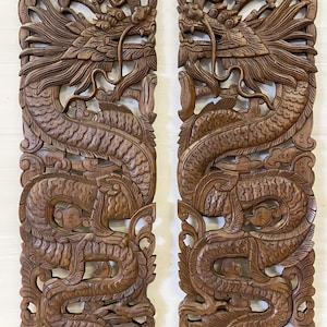 Könnte beinhalten: Zwei aufwendig geschnitzte Holzpaneele mit detaillierten Drachendesigns. Die Drachen sind mit fließenden Mähnen, Schuppen und gewundenen Körpern dargestellt, vor einem Hintergrund aus stilisierten Wolken und Blättern. Das Holz hat einen warmen, braunen Ton.