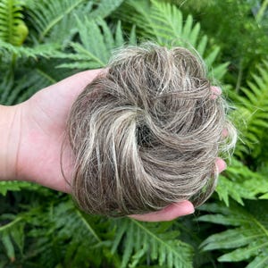 May include: A round hair bun extension featuring a blend of light brown, dark brown, and blonde highlights. The synthetic hair is styled in a messy, voluminous circular shape with varied strands creating a textured, multi-tonal appearance.