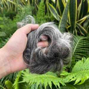 May include: A hair scrunchie featuring a mix of dark gray and silver-white synthetic hair fibers. The accessory has a voluminous, textured appearance with a circular shape, designed to be worn as a hair extension or bun wrap for styling.