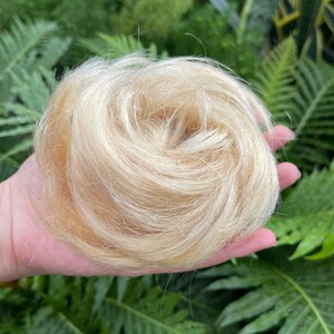 May include: A circular hair bun extension in a light blonde shade, featuring fine, straight synthetic fibers arranged in a swirled, layered pattern to create a voluminous, rounded shape that mimics the appearance of natural human hair.