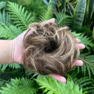 Op de afbeelding: Een ronde haarknot-extensie met een mix van donkerbruine en lichtblonde highlights. Het synthetische haar is gestyled in een rommelige, volumineuze cirkelvormige vorm met losse lokken, wat zorgt voor een getextureerd uiterlijk voor haarstyling.