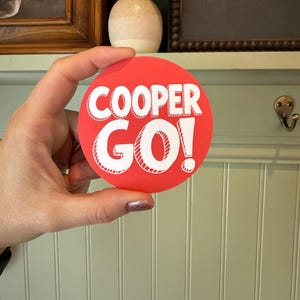 May include: A circular sticker with a vibrant red background featuring the white text COOPER GO! in a bold, sans-serif font. The letters have a subtle white outline and a slight shadow effect, creating a clean and legible graphic design on the sticker.