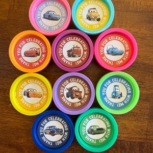May include: A collection of colorful, round party favors featuring cartoon car designs. Each favor has a different color rim and a central image of a car. Text around the car reads "YOU FOR CELEBRATING WITH ME! THANK".