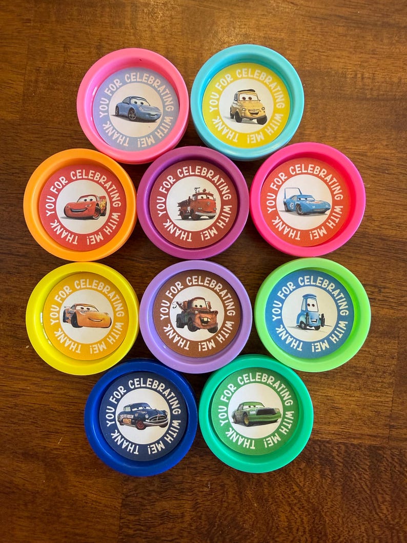 May include: A collection of colorful, round party favors featuring cartoon car designs. Each favor has a different color rim and a central image of a car. Text around the car reads "YOU FOR CELEBRATING WITH ME! THANK".