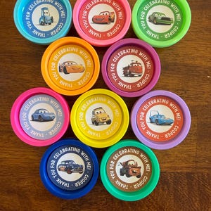 May include: A collection of colorful, round party favors featuring various cartoon car designs. Each favor has the text "Thank you for celebrating with me! Cooper" around the edge. The favors are in shades of blue, pink, green, orange, yellow, and purple.