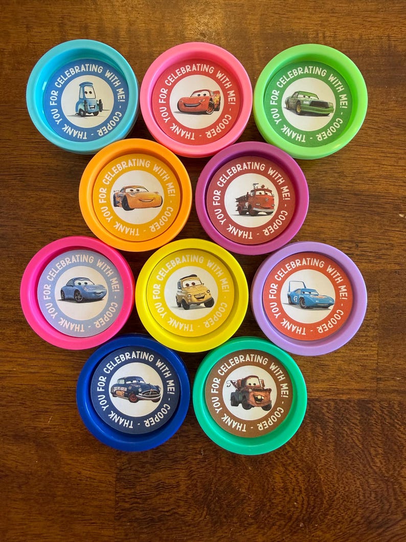 May include: A collection of colorful, round party favors featuring various cartoon car designs. Each favor has the text "Thank you for celebrating with me! Cooper" around the edge. The favors are in shades of blue, pink, green, orange, yellow, and purple.
