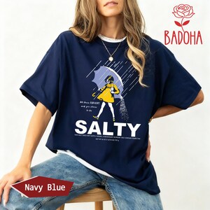 May include: Navy blue t-shirt with a graphic of a person under an umbrella, pouring salt. The word "SALTY" is printed in large white letters below the graphic. The shirt also has the text "All these flavors and you choose to be".