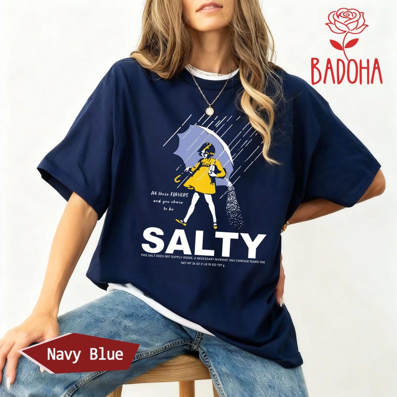 May include: Navy blue t-shirt with a graphic of a person under an umbrella, pouring salt. The word "SALTY" is printed in large white letters below the graphic. The shirt also has the text "All these flavors and you choose to be".