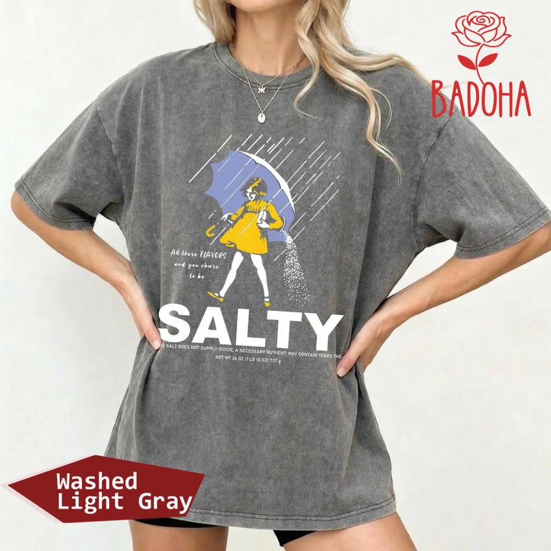 May include: Washed light gray t-shirt with a graphic of a person under a blue umbrella, with the word "SALTY" in large white letters. The shirt also has the text "All these flavors and you choose to be".
