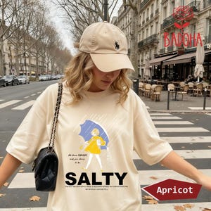 May include: Apricot-colored t-shirt with a graphic of a person under a blue umbrella, the word "SALTY" in large letters, and the text "All these flavors and you choose to be." A beige baseball cap with a black logo is also visible.
