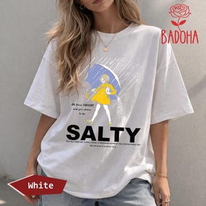 May include: White t-shirt with a graphic of a person under a blue umbrella in the rain, with the word "SALTY" in large letters. The shirt also has the text "All these flavors and you choose to be".
