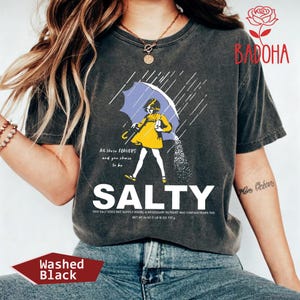 May include: Washed black t-shirt with a graphic of a person under a blue umbrella, with the word "SALTY" in large white letters. The shirt also has the text "At those flavors and you choose to be".