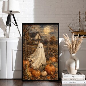 May include: A framed rectangular wall art print featuring a white ghost standing in a pumpkin patch at night. The scene includes a rustic house, a wooden fence, and a full moon in the background. The artwork uses warm orange, brown, and dark tones.