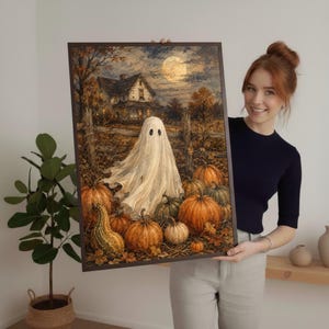 May include: A framed rectangular wall art print featuring a white ghost standing in a pumpkin patch in front of a house under a full moon. The scene includes orange pumpkins, a wooden fence, and autumn trees in a dark, atmospheric color palette.