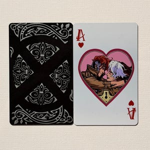Könnte beinhalten: Spielkarte mit dem Herz-Ass. Die Vorderseite der Karte zeigt ein Paar in einem rosa Herzen, und die Rückseite ist schwarz mit einem silbernen, verzierten Design. Das Herz-Ass-Symbol befindet sich in den oberen linken und unteren rechten Ecken.