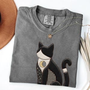 Peut inclure: T-shirt gris avec un appliqué de chat noir et blanc orné d'un croissant de lune. Le t-shirt a un col rond et des manches courtes. Un collier argenté avec un petit pendentif repose sur le t-shirt. L'étiquette indique "Comfort Colors".