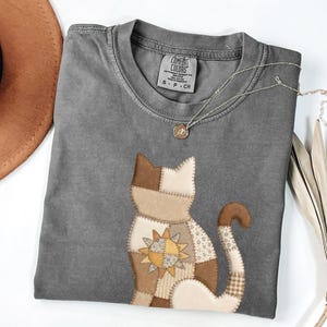 May include: A grey Comfort Colors t-shirt with a patchwork cat design in brown and cream tones. The cat has a sunburst design. A silver necklace with a small pendant is on the shirt. A brown hat is visible in the upper left corner.