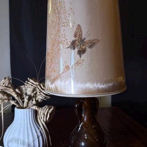 Op de afbeelding: Een vintage tafellamp met een bruine keramische voet en een beige lampenkap. De lampenkap heeft een vlinder- en bloemenontwerp. Een witte vaas met gedroogde planten staat in de buurt.