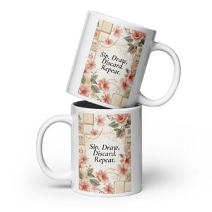 Könnte beinhalten: Zwei weiße Keramikbecher, die übereinander gestapelt sind. Jeder Becher hat den Text "Sip. Draw. Discard. Repeat." mit floralen und Kachelmustern. Die Becher haben einen gebogenen Griff.