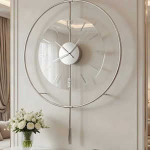 Pode incluir: Um relógio de parede moderno e minimalista com um mostrador de vidro transparente e uma moldura de metal prateado. O relógio apresenta um design circular com marcadores de hora e ponteiros simples. Está montado numa parede clara, com um buquê de flores brancas por baixo.