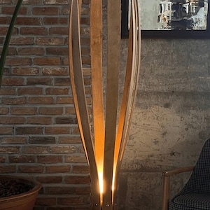 May include: A tall, wooden floor lamp with a unique design. The lamp features multiple vertical wooden slats that curve inward, creating an open, organic shape. Warm light emanates from within, illuminating the surrounding area. The lamp is set against a brick wall.