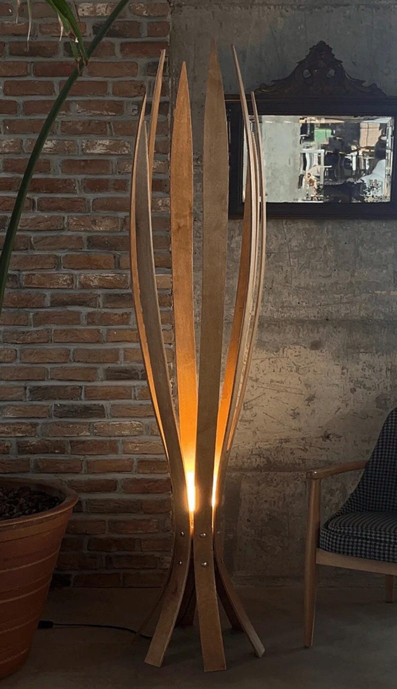 May include: A tall, wooden floor lamp with a unique design. The lamp features multiple vertical wooden slats that curve inward, creating an open, organic shape. Warm light emanates from within, illuminating the surrounding area. The lamp is set against a brick wall.
