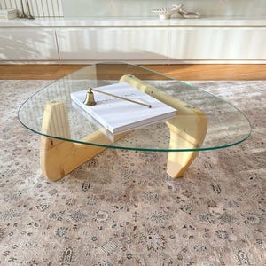 May include: A modern, triangular glass coffee table with light wood legs. A book with the word "BARCELONA" is on the table, along with a small brass bell. The table sits on a patterned rug.