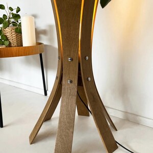 May include: A wooden floor lamp with a unique, curved design. The lamp has a light brown finish and is held together with silver-colored screws. The lamp's legs are angled outward, and a black power cord extends from the base.