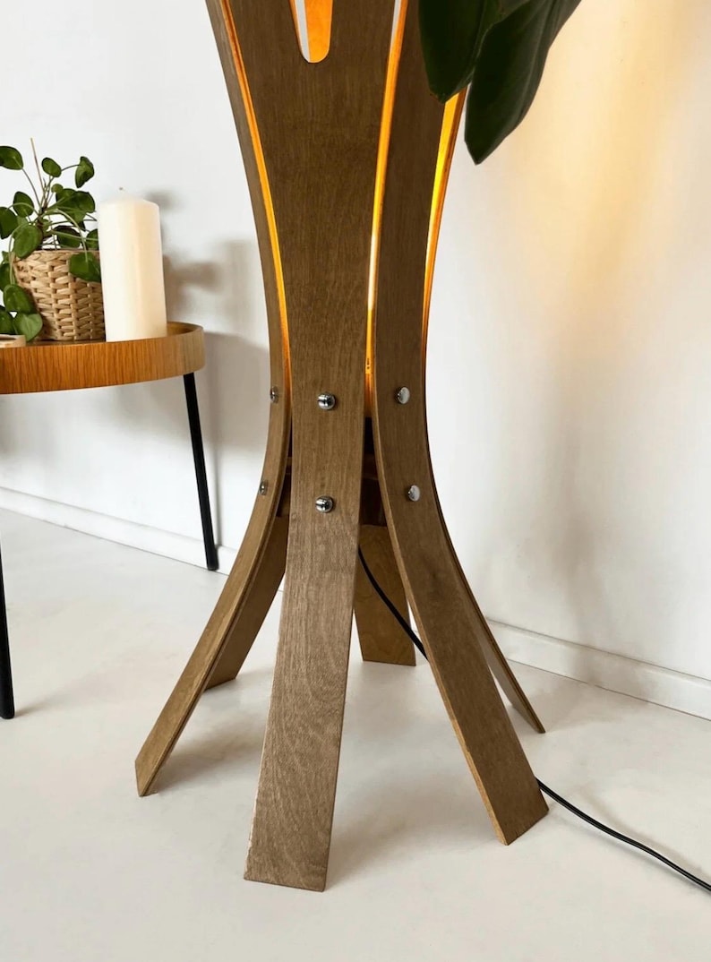 May include: A wooden floor lamp with a unique, curved design. The lamp has a light brown finish and is held together with silver-colored screws. The lamp's legs are angled outward, and a black power cord extends from the base.