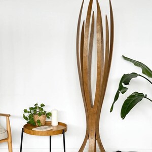 May include: A tall, wooden floor lamp with a unique, open design. The lamp features multiple vertical wooden slats that curve upwards, creating a sculptural, organic form. The lamp is made of light brown wood and stands on a base with multiple legs.