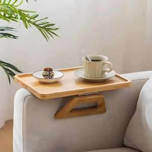 Puede incluir: Bandeja de brazo de sofá de bambú con una taza de café y un pequeño postre. La bandeja está sujeta al brazo de un sofá de color claro. La bandeja es de madera clara y mide unos 30 cm de largo.