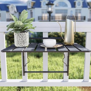 May include: A small, dark gray wooden balcony table with a metal frame is attached to a white railing. A white ceramic cup and saucer with a cookie sit on the table, along with a gold watering can and a potted plant.