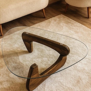 Könnte beinhalten: Moderner, dreieckiger Couchtisch mit klarer Glasplatte und dunklem Holzfuß. Der Fuß hat ein einzigartiges, skulpturales Design. Der Tisch steht auf einem hellen Teppich, im Hintergrund zwei beige Sessel.