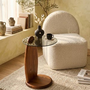 Puede incluir: Una mesa auxiliar moderna con una tapa redonda de cristal y una base de madera curvada. La mesa está junto a una silla redondeada de color crema. Una taza de café negra está sobre la mesa, junto con un pequeño objeto de madera y un jarrón negro con tallos decorativos.