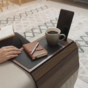 Könnte beinhalten: Ein dunkles Sofa-Armtablett aus Holz mit schwarzer Oberfläche, das eine Kaffeetasse, ein Smartphone, ein braunes Leder-Notizbuch und einen Stift hält. Das Tablett ist so konzipiert, dass es über die Armlehne eines Sofas passt.