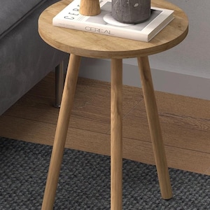 Pode incluir: Pequena mesa lateral redonda de madeira clara com três pernas anguladas. No tampo, um vaso de madeira, um recipiente cinza com efeito mármore e um livro intitulado "Cereal". A mesa está sobre um tapete cinza.