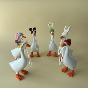 May include: A group of white goose figurines with orange beaks and feet, each wearing a different crocheted hat. Hats include a pink hat with a pom-pom, a bear hat, a flower hat, a bunny hat, a red hat, and a rainbow hat. The geese have white strings attached to their beaks.