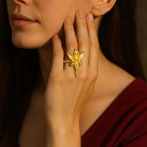 Puede incluir: Un anillo dorado con un diseño detallado de hada. El anillo presenta un hada esculpida con alas extendidas y un vestido fluido. El anillo se lleva en un dedo, mostrando sus intrincados detalles. El anillo es una pieza de joyería.