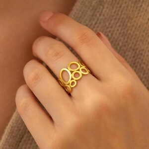Op de afbeelding: Een goudkleurige ring met een open ontwerp van onderling verbonden cirkels en ovalen. De ring wordt aan een vinger gedragen en toont het ingewikkelde, geometrische patroon. Het sieraad heeft een gepolijste afwerking die licht reflecteert.