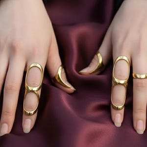 May include: Gold-toned rings on display. The image showcases a hand adorned with several gold rings, including a double-finger ring and simple bands. The rings are set against a deep burgundy fabric, creating a contrast that highlights the jewelry.