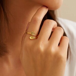 May include: A gold-colored ring featuring a fork and spoon design. The ring wraps around the finger, with the fork and spoon positioned on opposite sides. The ring is a unique piece of jewelry.
